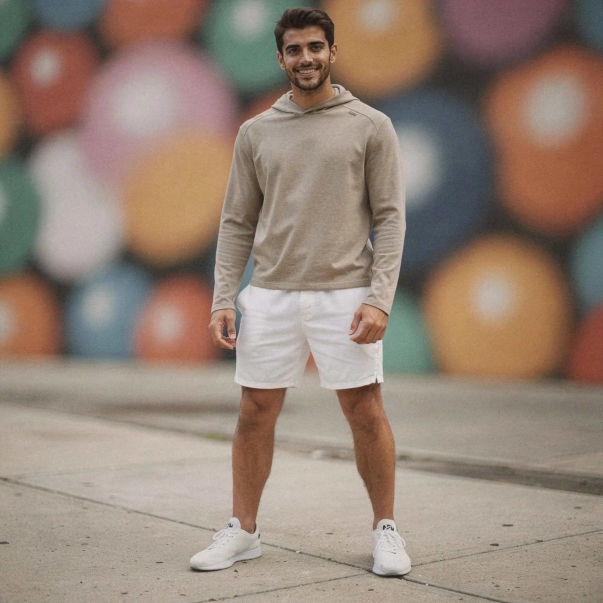 Man wearing beige slim fit hoodie and white shorts, standing outdoors, colorful mural in background