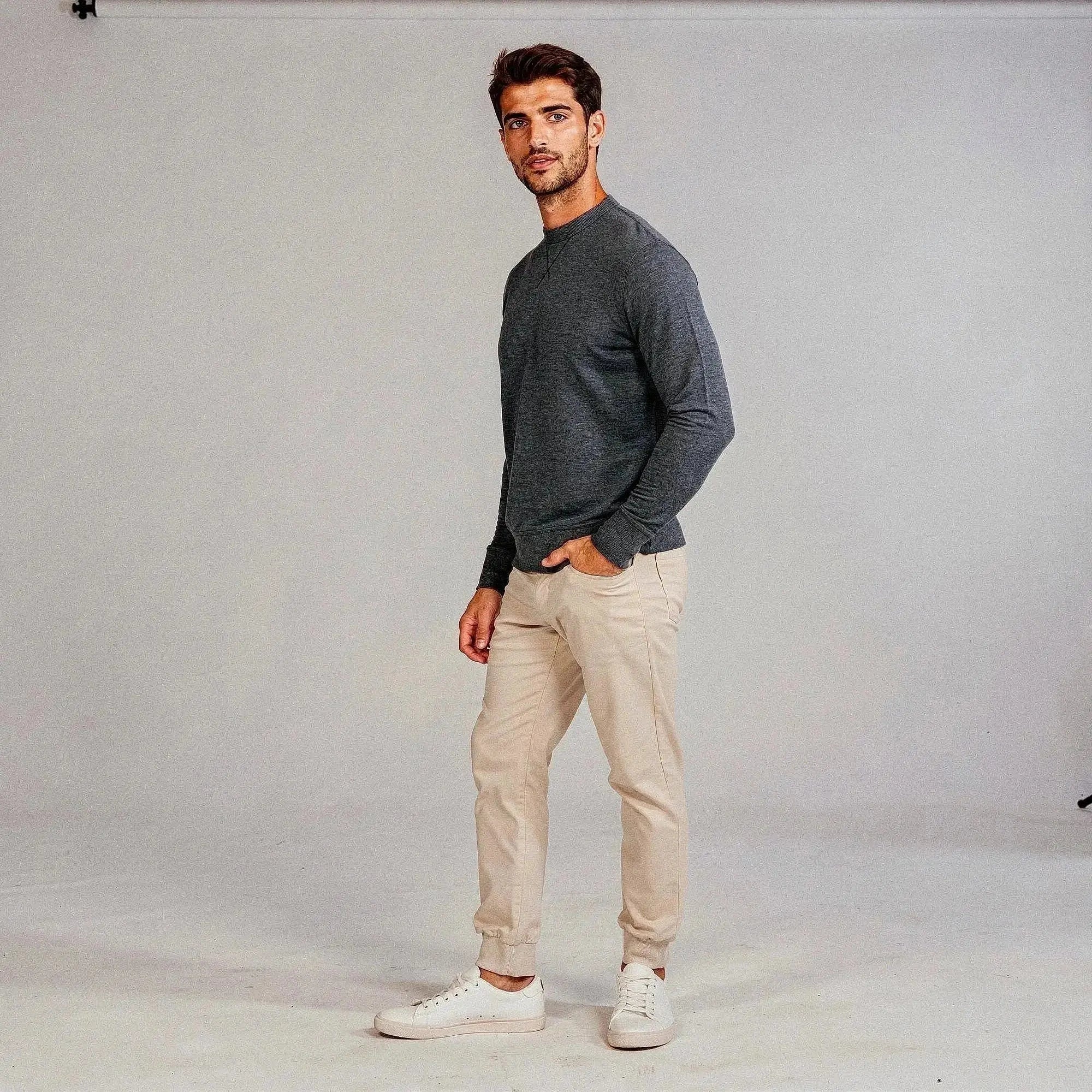 Man wearing a dark gray long sleeve sweatshirt and beige pants, standing sideways in a neutral