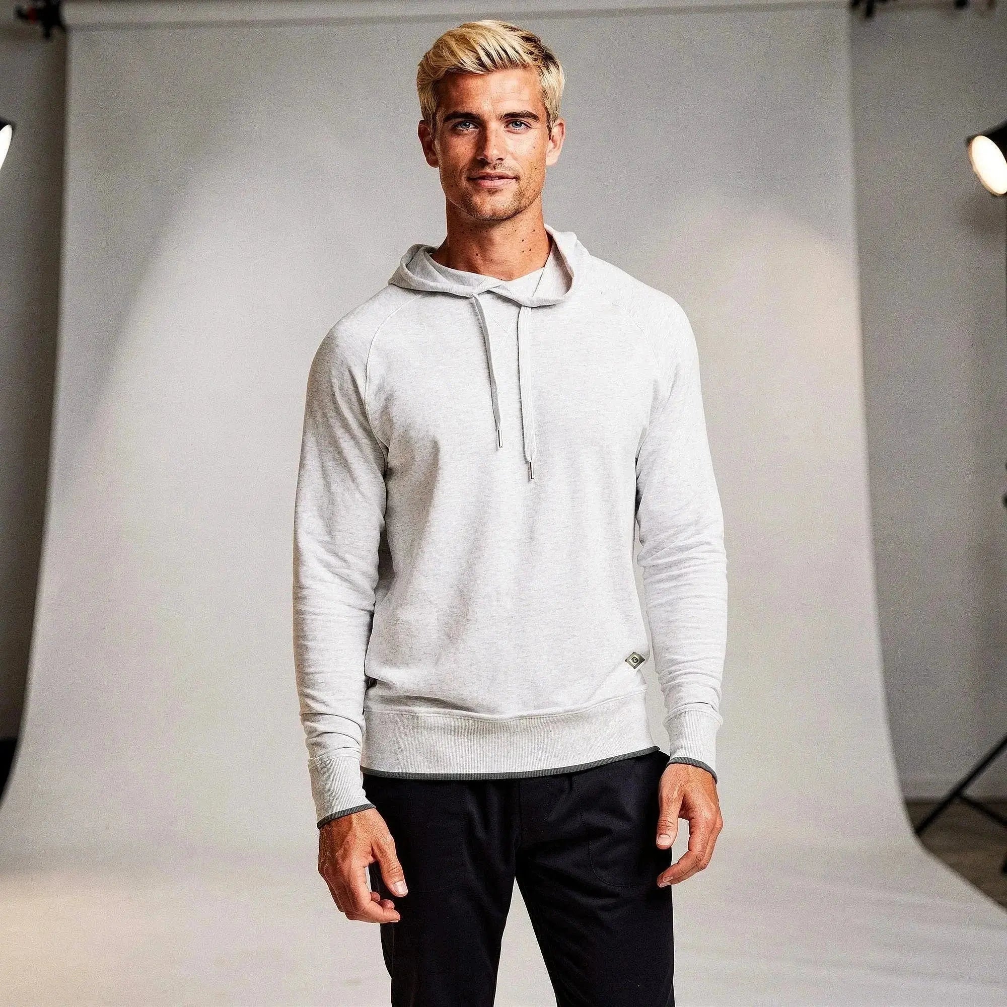 Man wearing a light gray long sleeve hoodie, standing confidently with hands relaxed, against a backdrop
