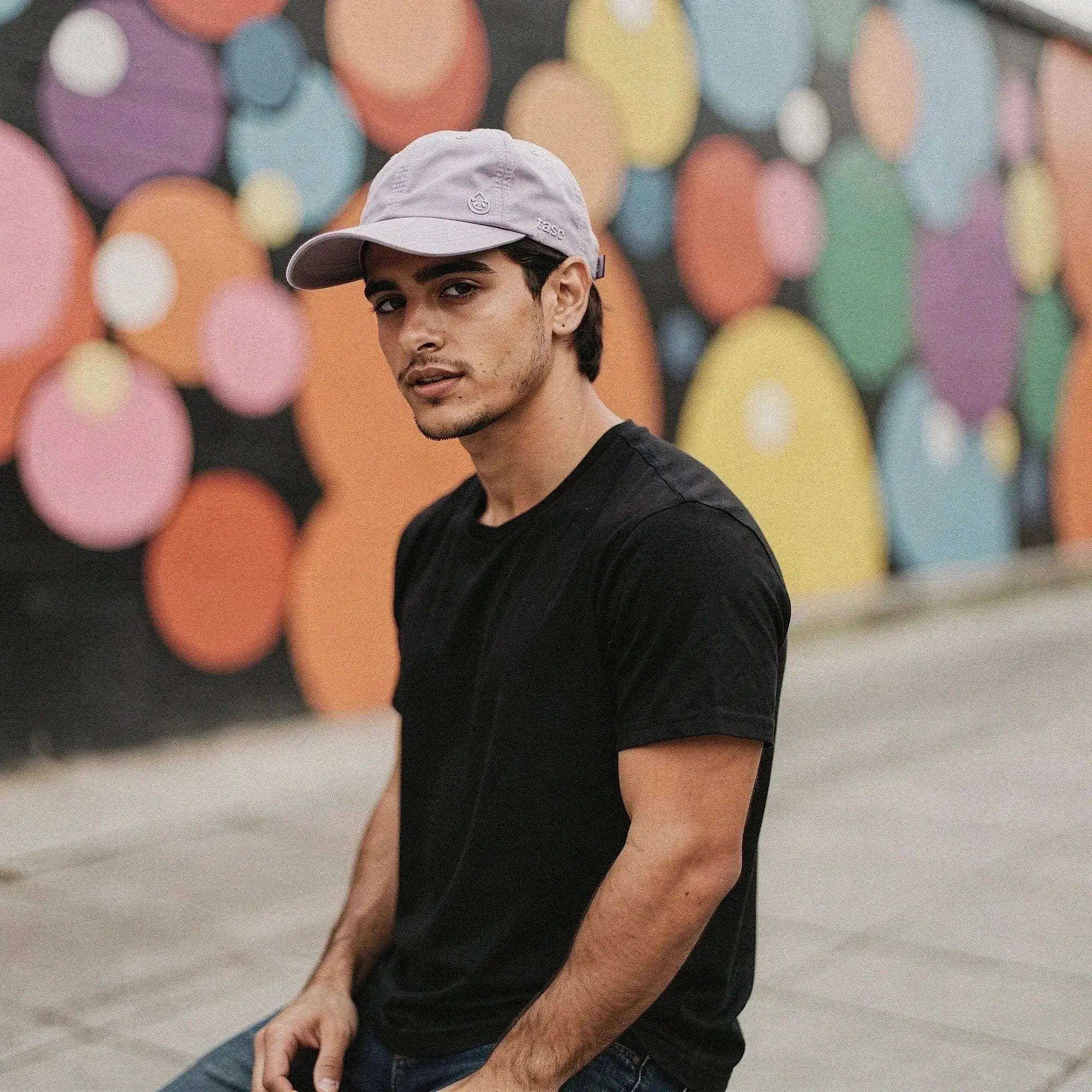 Man wearing light purple cap and black t shirt stands in of colorful circle mural
