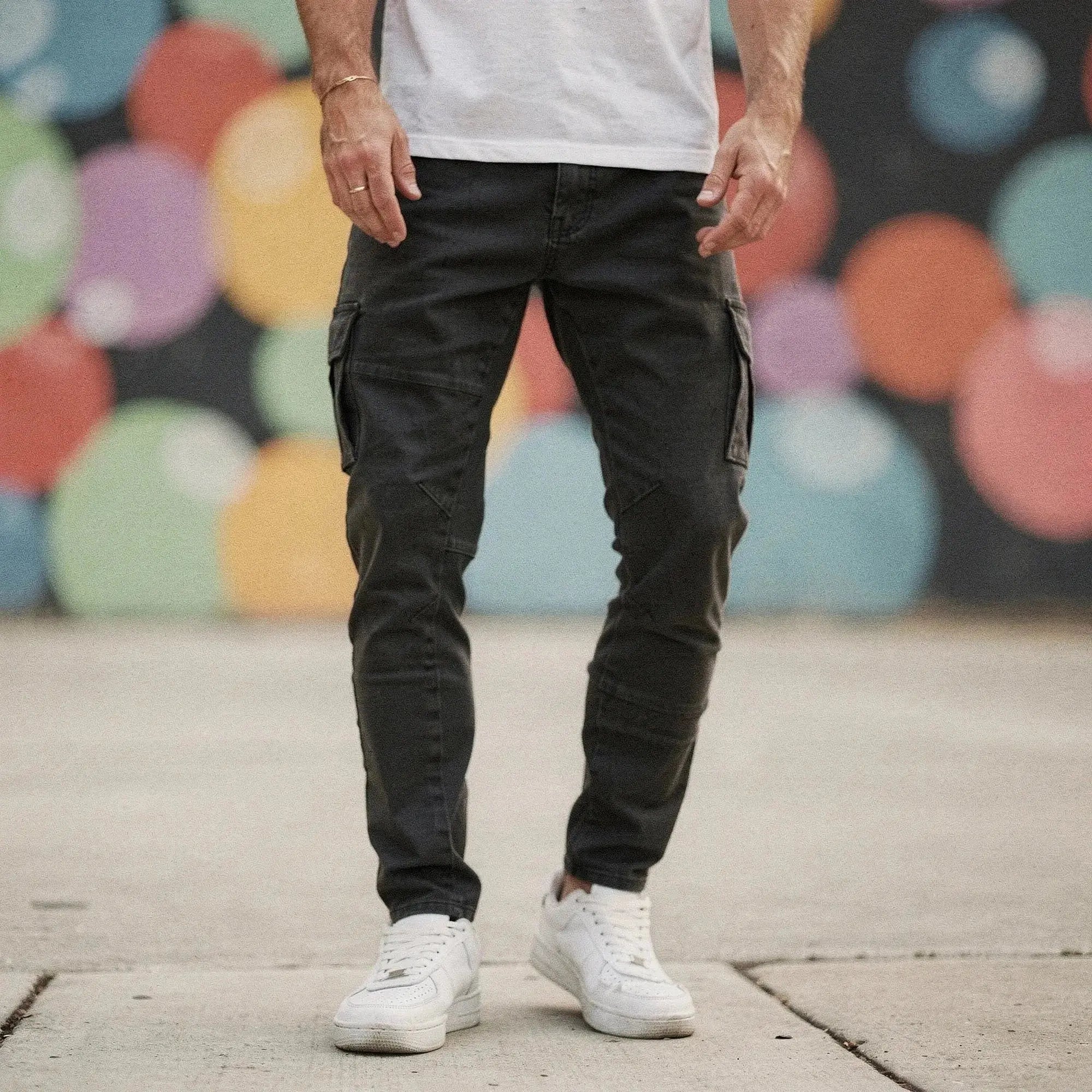 Man wearing dark grey slim fit cargo trousers with pockets, standing on pavement, colorful wall behind
