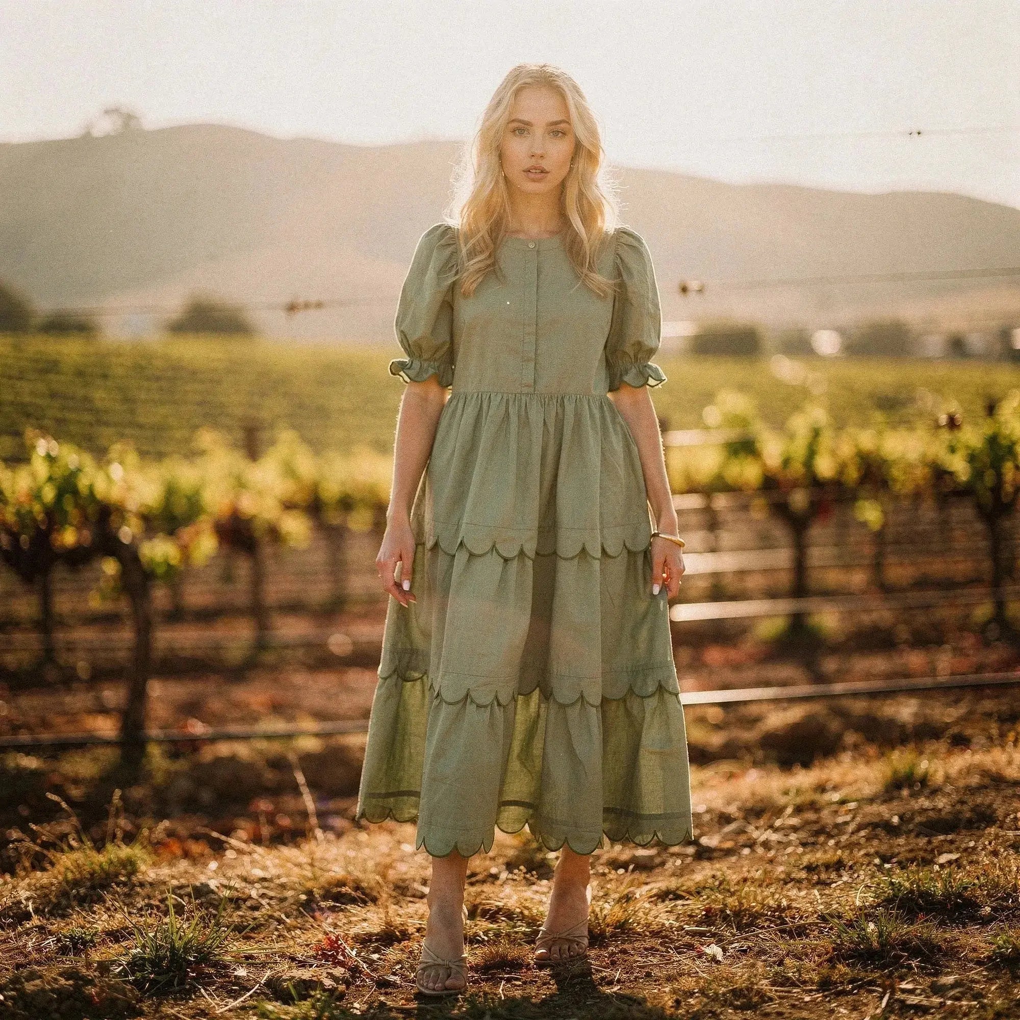 Woman wearing a sage green flowy A line midi dress with scalloped hem, standing in a vineyard
