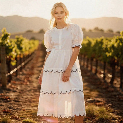 Woman wearing a white A line midi dress with scalloped edges, standing in a vineyard at sunset