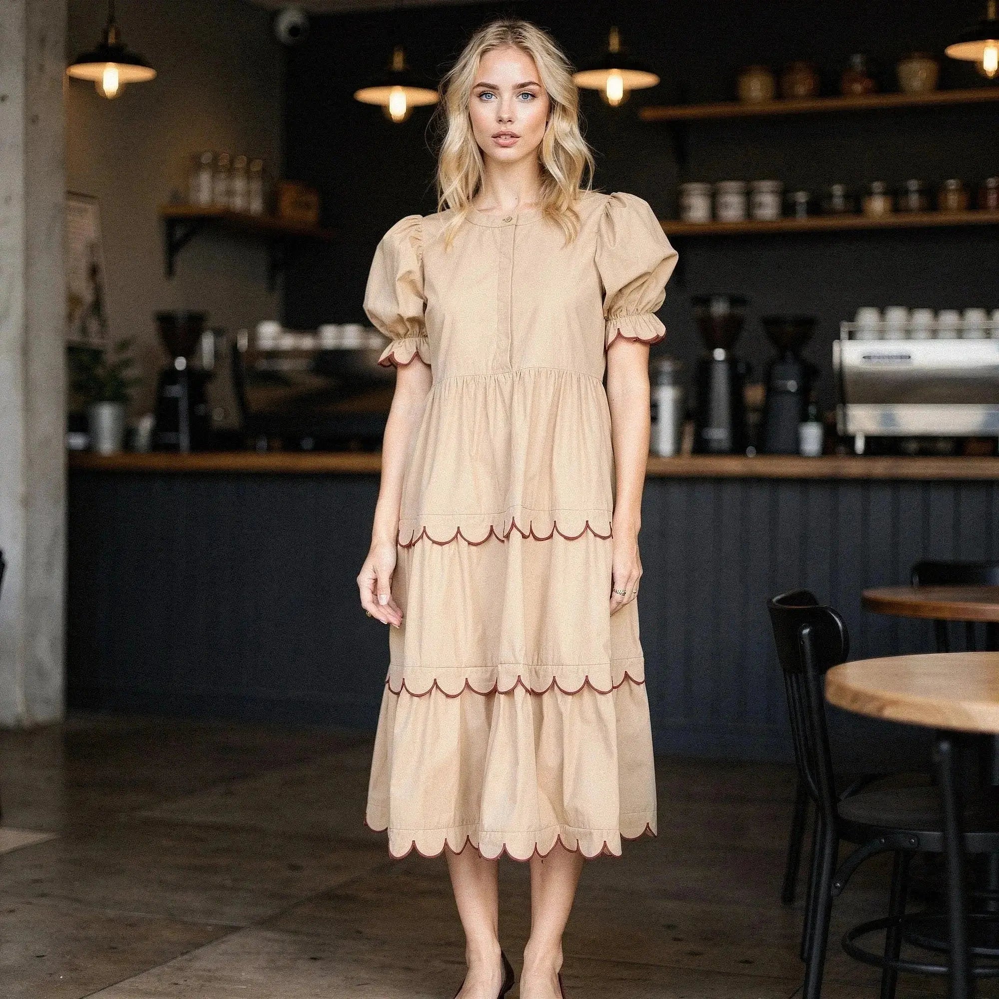 Woman wearing beige A line midi dress with scalloped hem, standing in a cafe setting