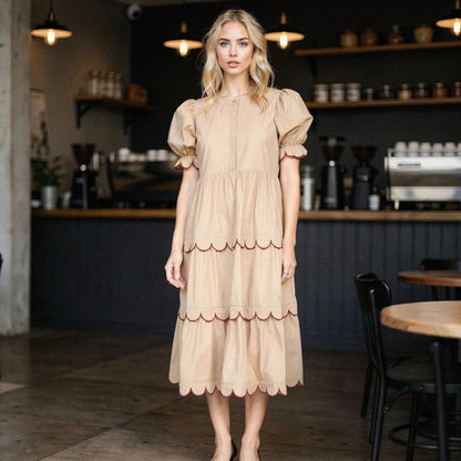 Woman wearing beige A line midi dress with scalloped hem, standing in a cafe setting