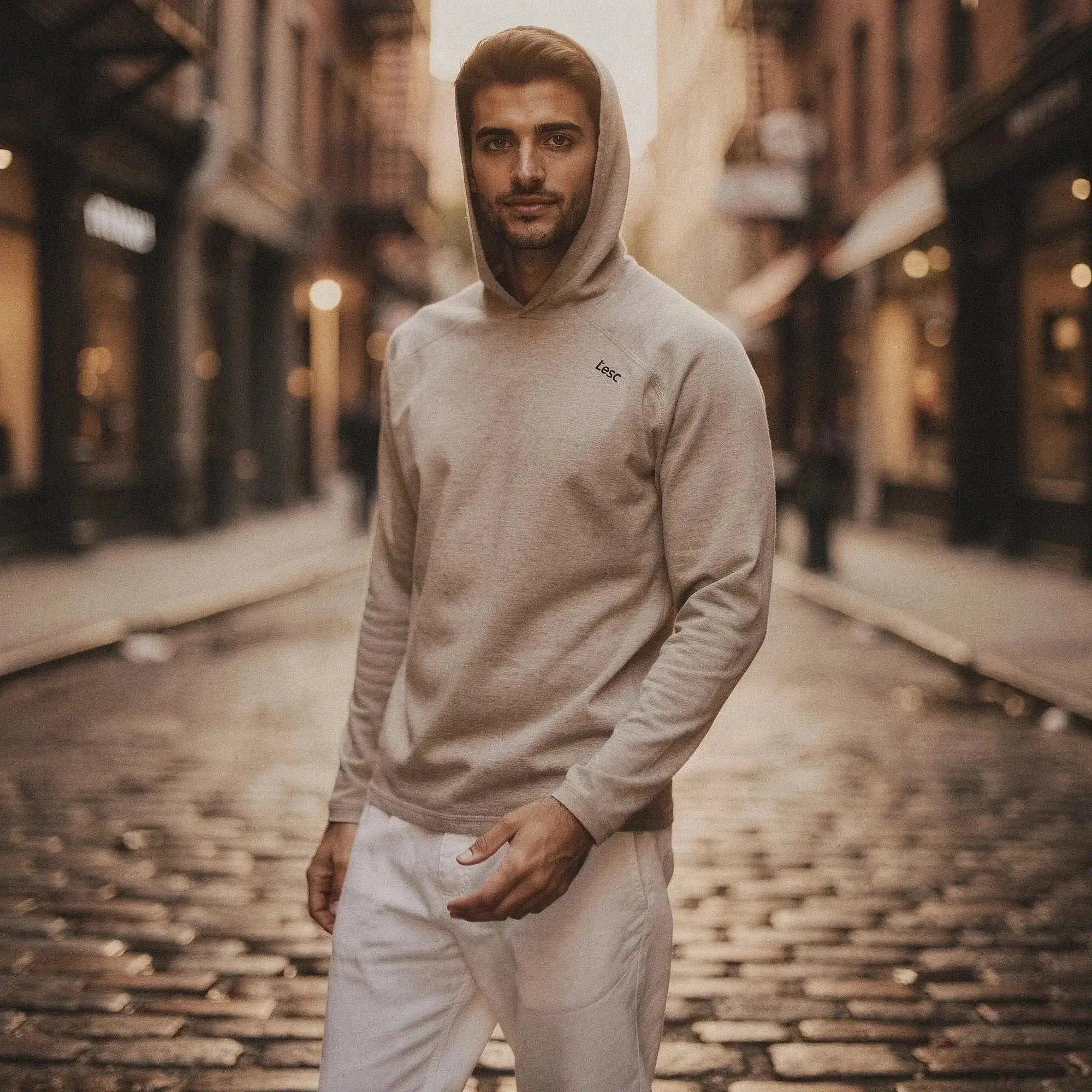 Man wearing a beige slim fit hoodie with hood up, standing on cobblestone street, hands