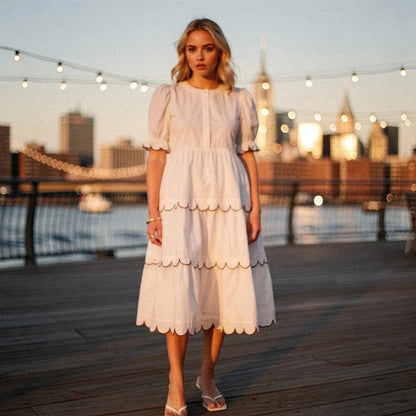 Woman wearing a white A line midi dress with scalloped edges, standing on a boardwalk at sunset