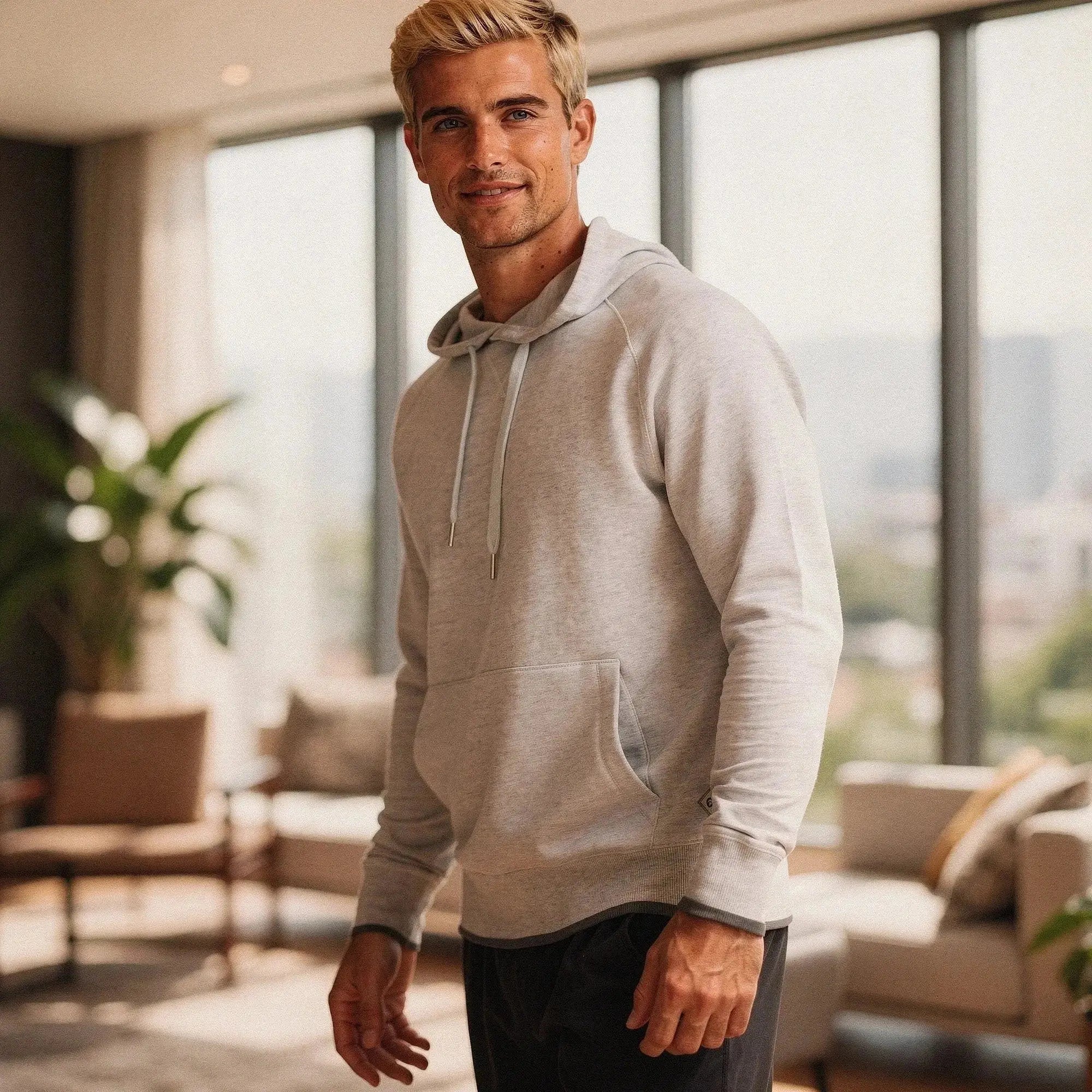 Man wearing a light gray long sleeve hoodie with drawstrings, standing indoors near large windows