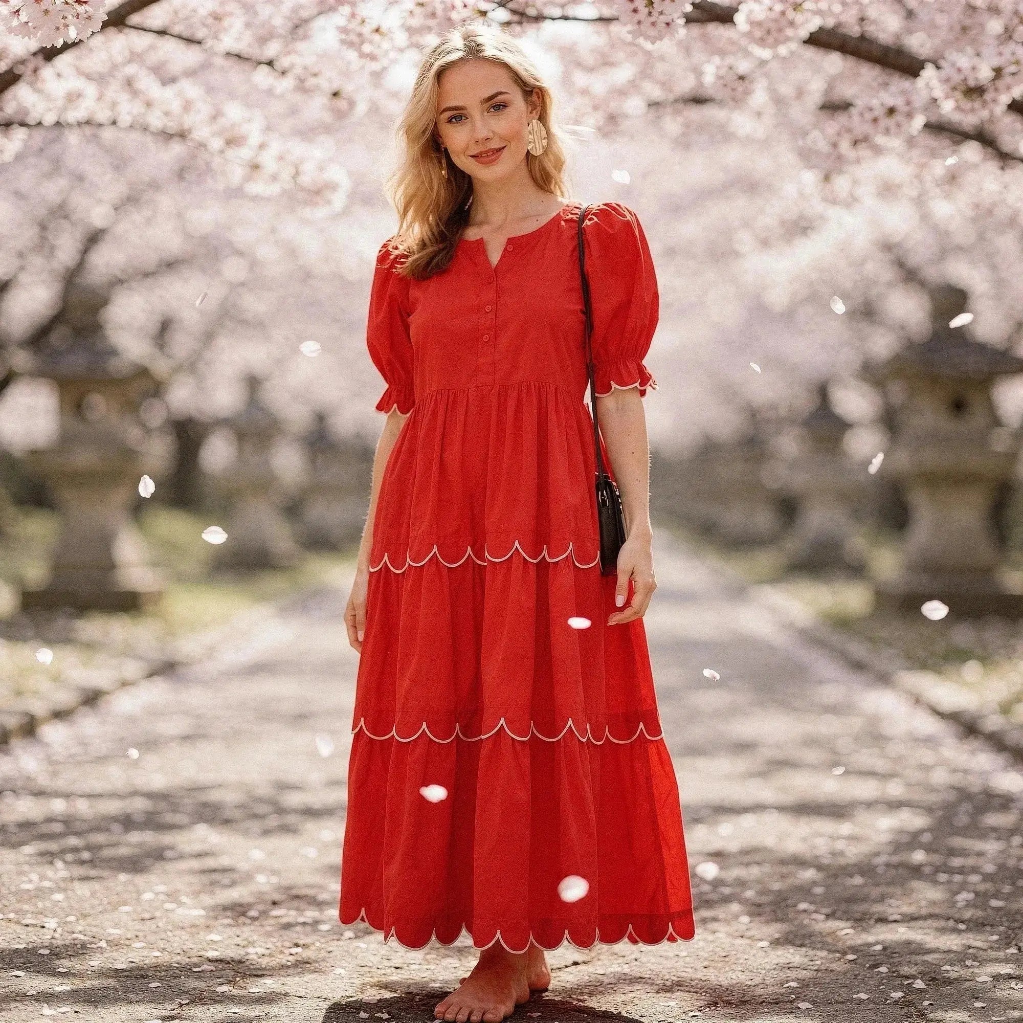 Woman wearing a red A line midi dress with scalloped hem, standing barefoot under cherry blossoms