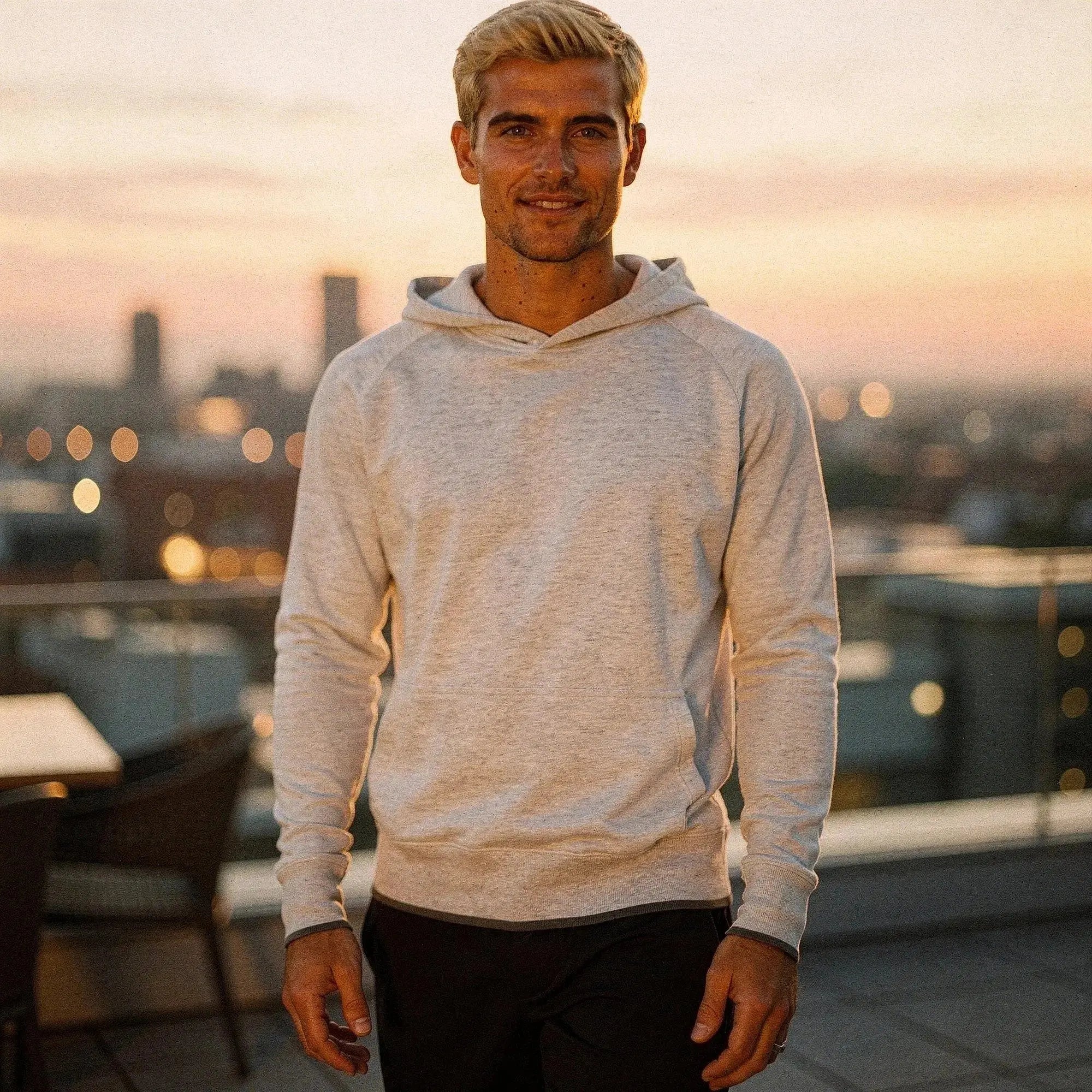 Man wearing a light gray long sleeve hoodie, standing outdoors at sunset, with city skyline background
