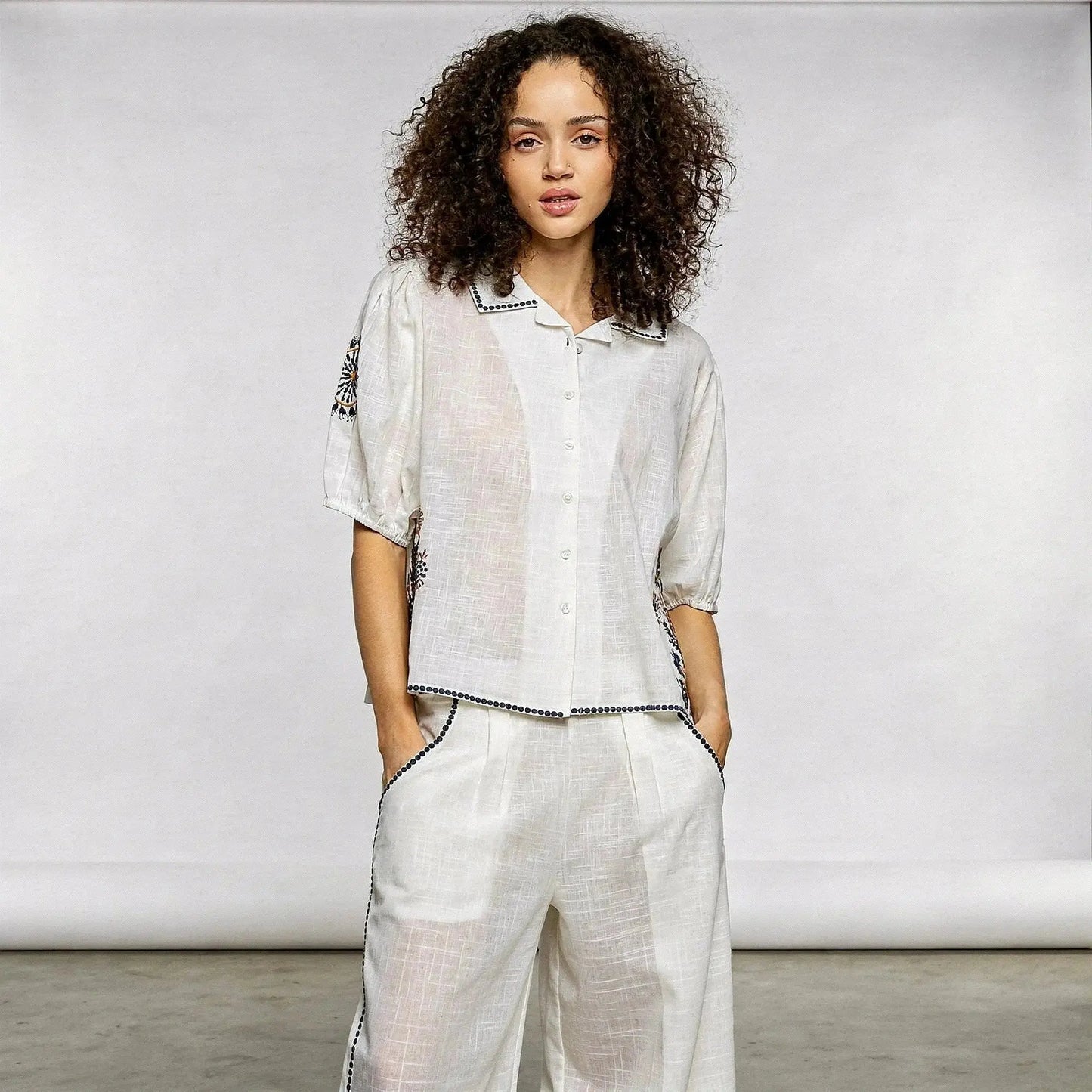 Woman wearing a white short sleeve shirt with floral embroidery, hands in pockets, standing confidently
