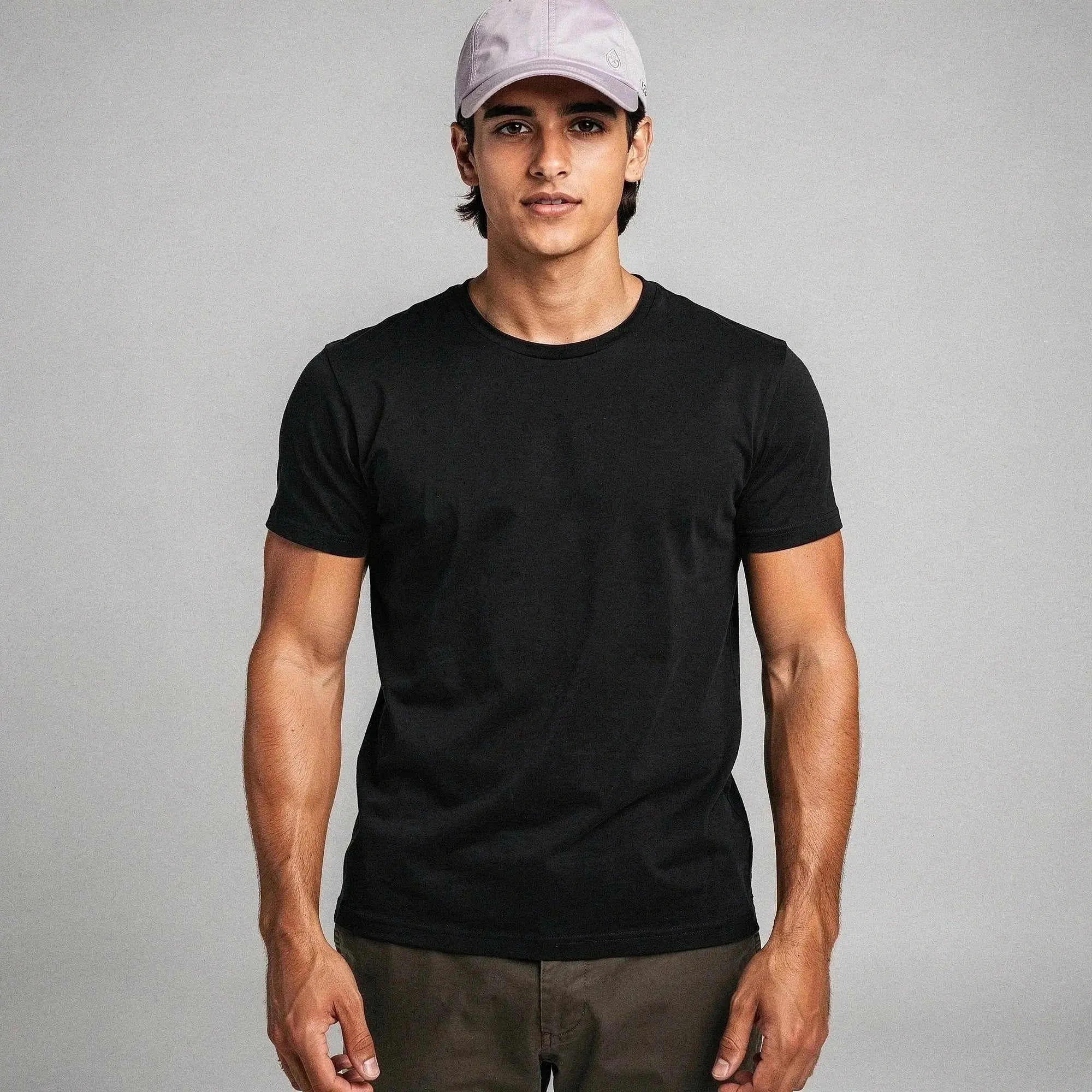 Man wearing a light gray cap, black t shirt, and brown pants, standing against a plain background