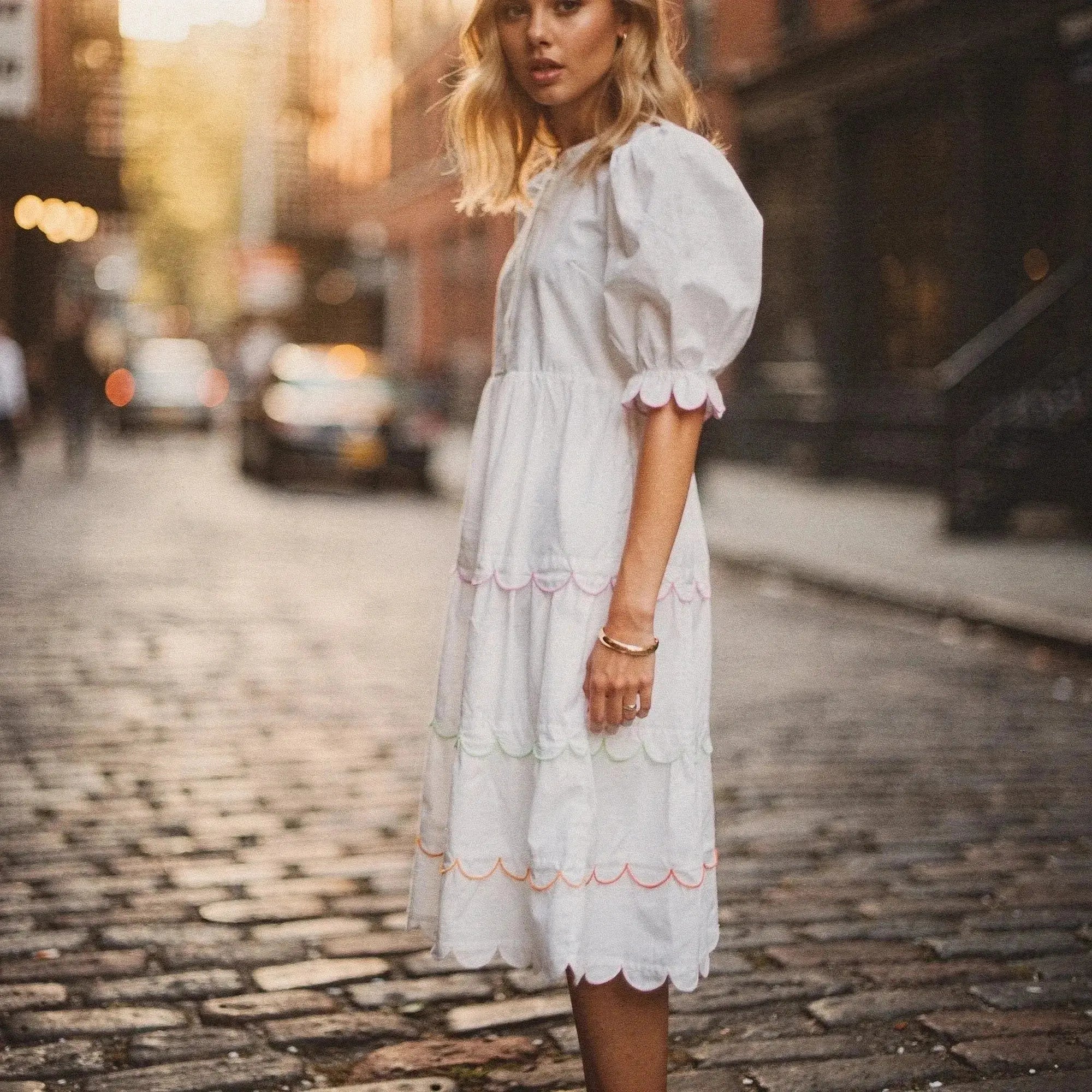 Woman wearing a white flowy A line midi dress with scalloped edges, standing on a cobblestone street